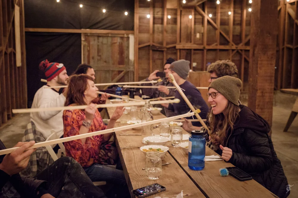 Participants sharing a playful meal with long utensils at a wooden table